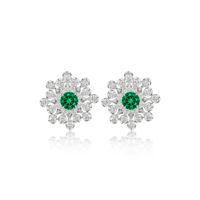 The Gem Vivi Emerald Snowflake Stud Earrings feature a central green gemstone surrounded by sparkling clear stones in a flower shape, all set in 18k white gold.