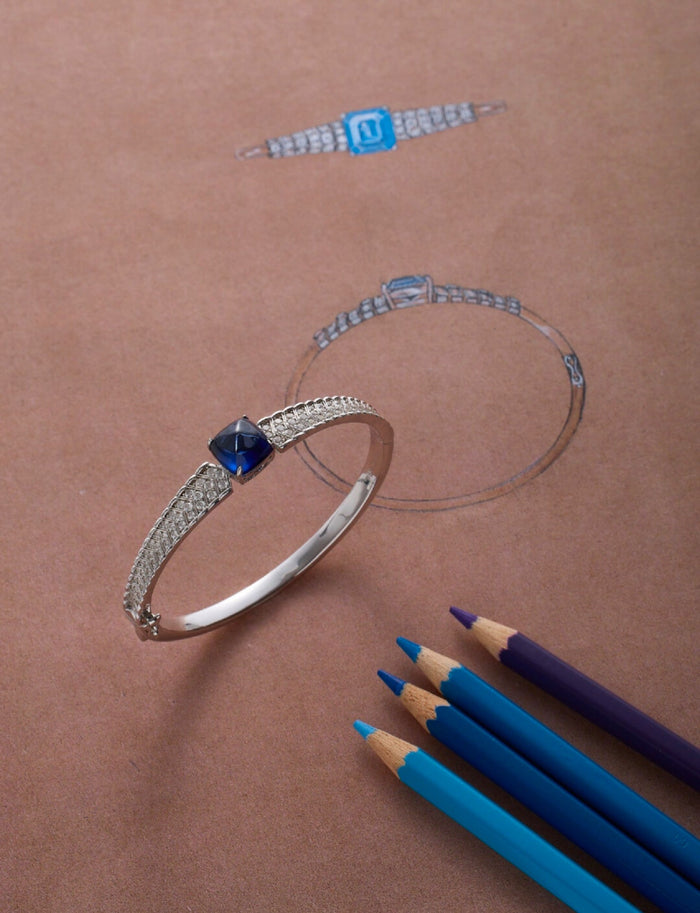 The Gem Vivi Blue Cabochon Bracelet is displayed on brown paper with its sketch, beside blue and purple colored pencils.