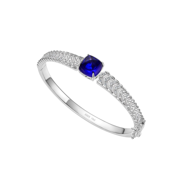 The Gem Vivi Blue Cabochon Bracelet showcases a blue gemstone centerpiece and a row of clear stones on a sterling silver band, elegantly displayed on a white background—perfect for any refined occasion.